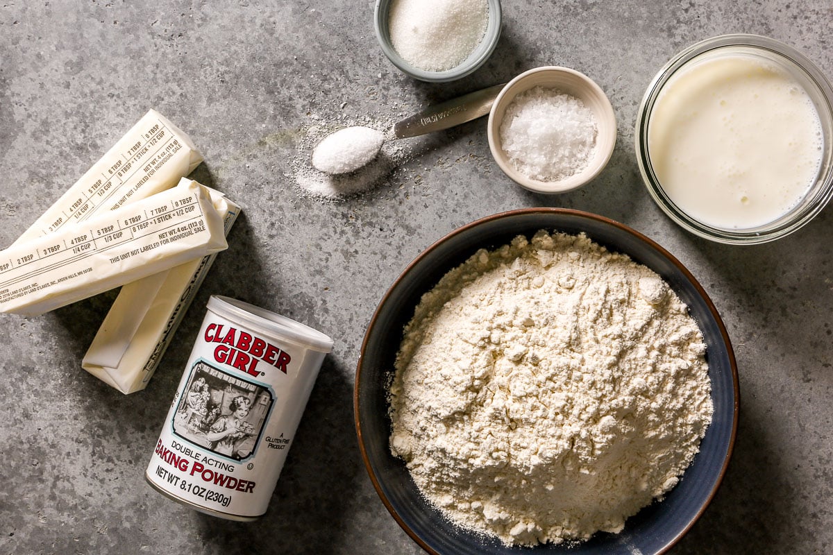 Butter, baking powder, flour, salt, sugar, and buttermilk milk are arranged on a gray countertop.