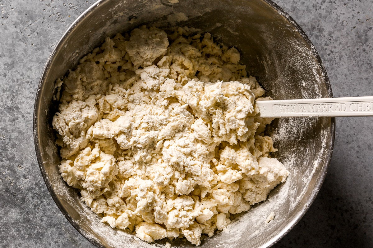 A metal bowl with biscuit dough being mixed using a white rubber spatula on a gray surface.