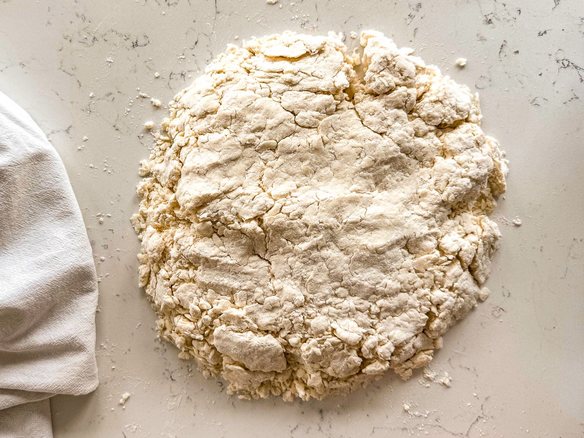 Rough biscuit dough mound on a floured white countertop, with a kitchen towel on the left side.