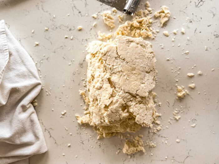 Shaggy biscuit dough folded onto itself in thirds on a white countertop, with a white cloth and metal dough cutter nearby.