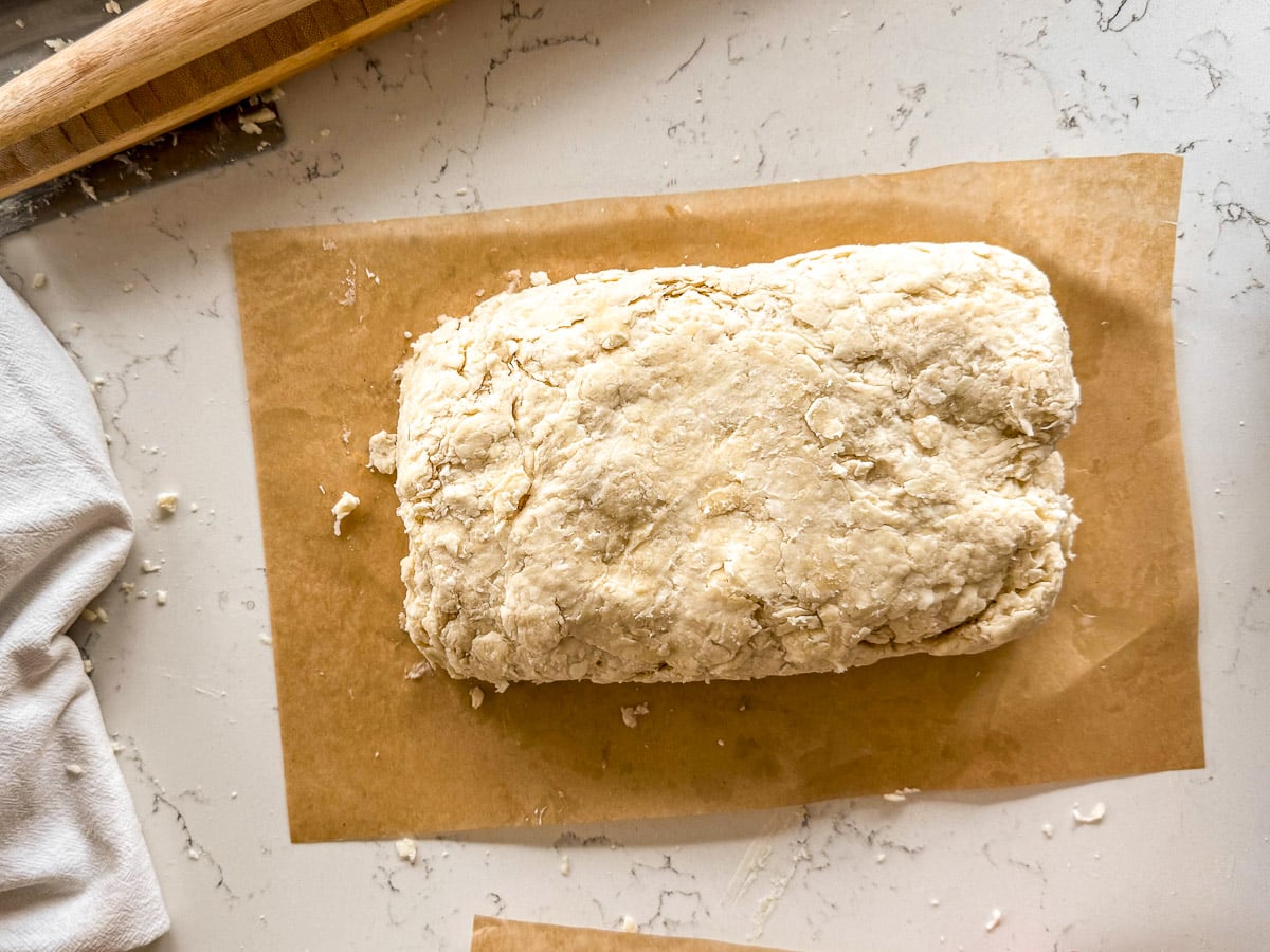 A rectangular block of dough sits on parchment paper on a light marble countertop with a rolling pin nearby.