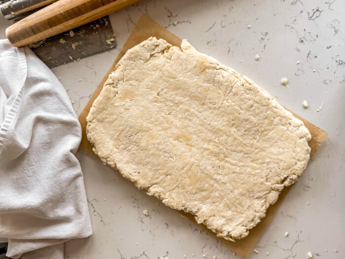 Rectangular slab of biscuit dough on parchment with a rolling pin and towel nearby on a flour-dusted counter.