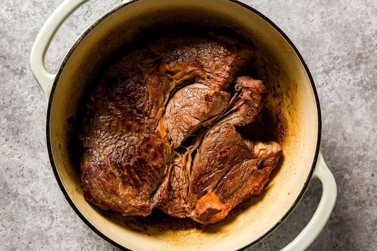 Cooked chuck roast with browned edges in a round, light-colored Dutch oven on a gray surface.