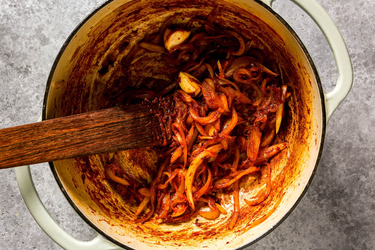 Sautéed onions, tomato paste, and spices in a pot being stirred with a wooden spatula on a gray surface.