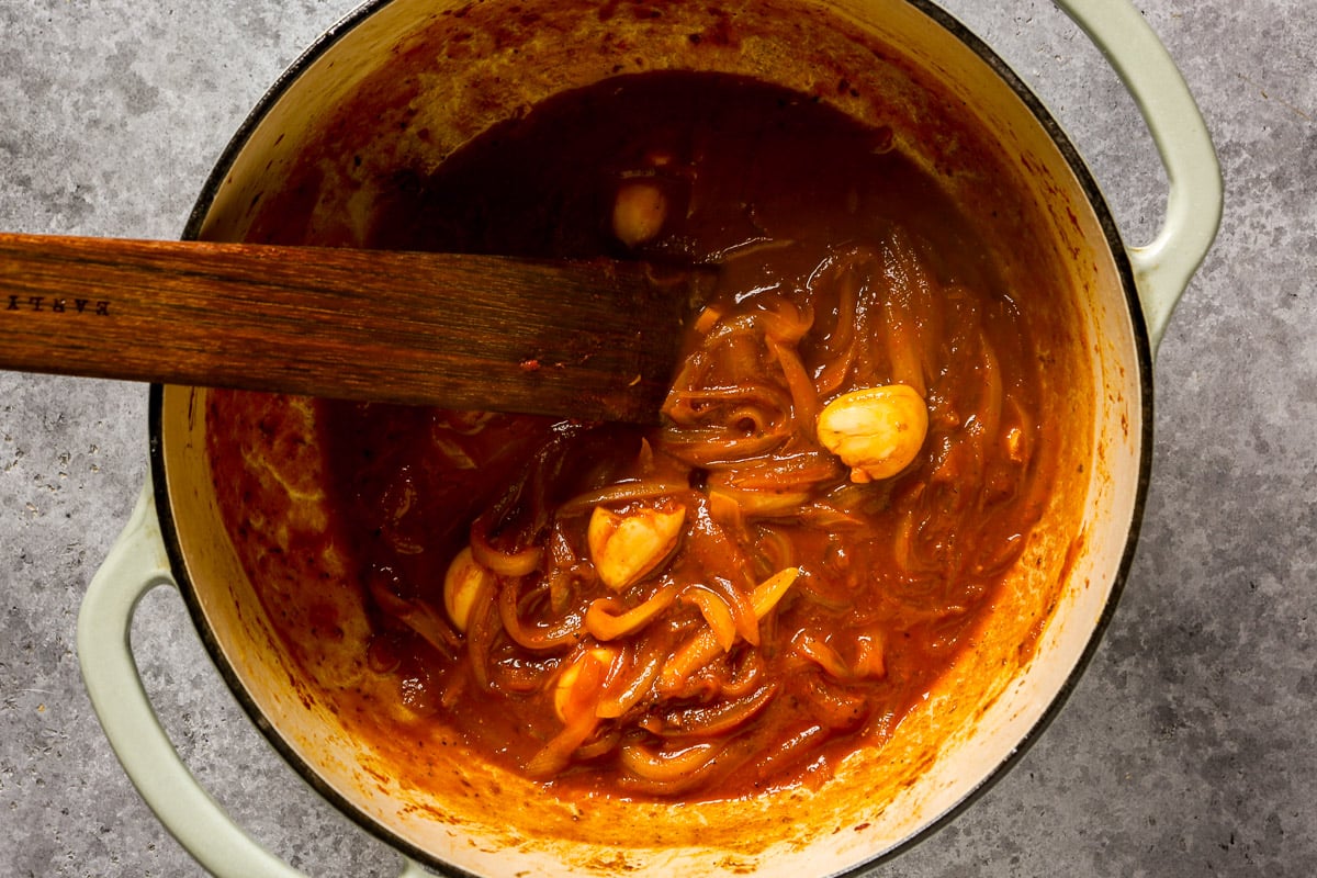 Sliced onions and garlic cooking in a red tomato sauce in a large pot, stirred with a wooden spatula.