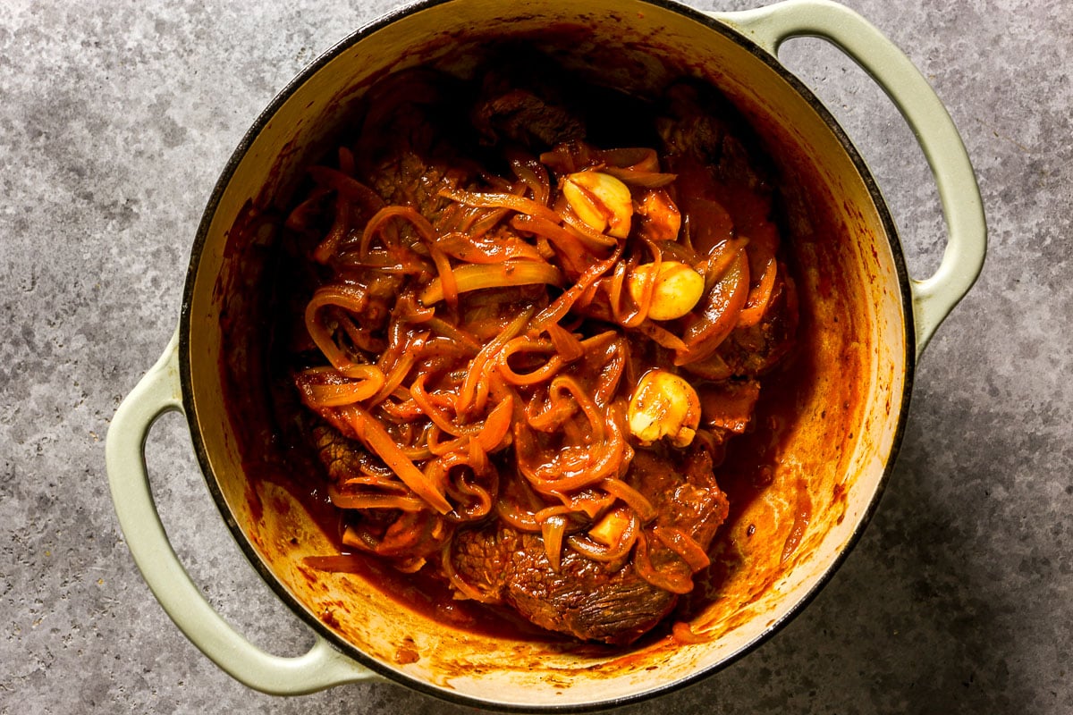 Beef chuck roast with onions and whole garlic cloves in a red sauce, cooked in a light green Dutch oven.