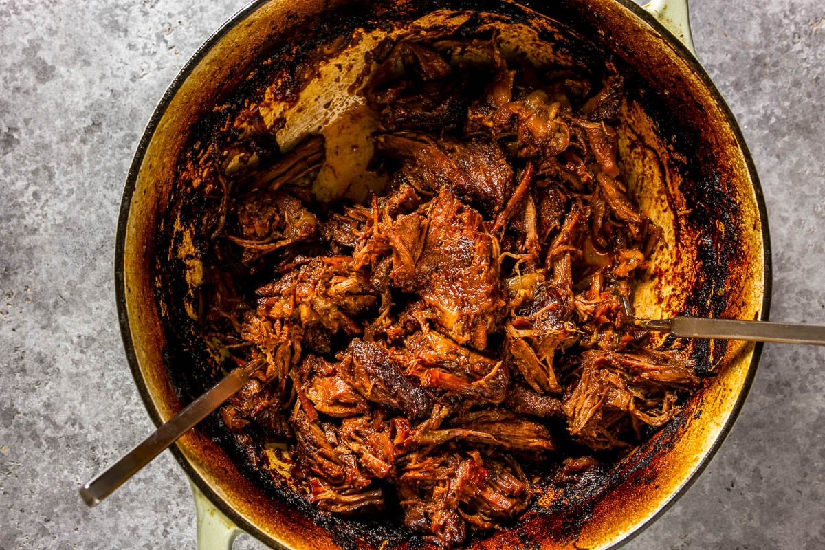 Shredded, cooked beef in a pot with two metal utensils on a gray surface.