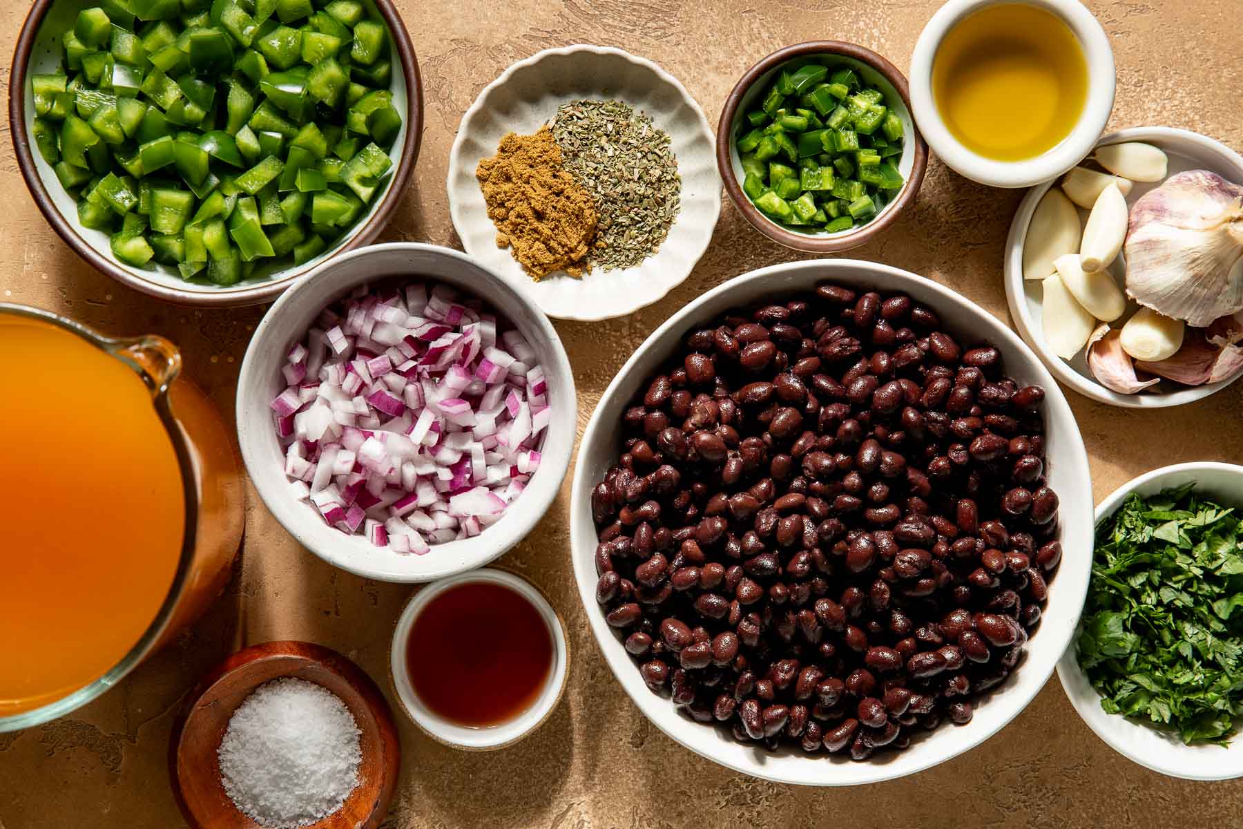 Bowls of black beans, diced green peppers, red onion, cilantro, jalapeño, garlic, cumin, dried oregano, oil, broth, and salt on a tabletop.
