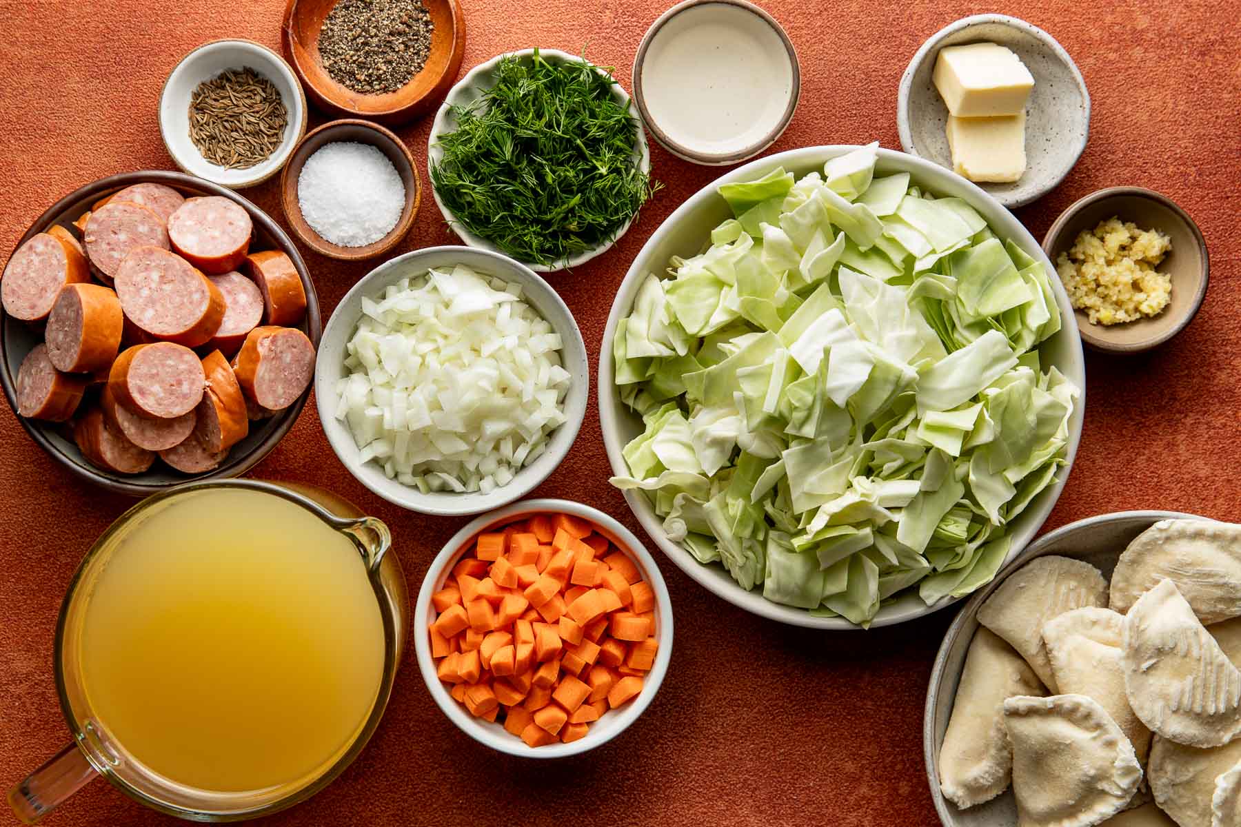 Bowls of chopped cabbage, carrots, onions, sliced sausage, pierogi, broth, butter, dill, caraway, and salt set out on a rusty orange counter.