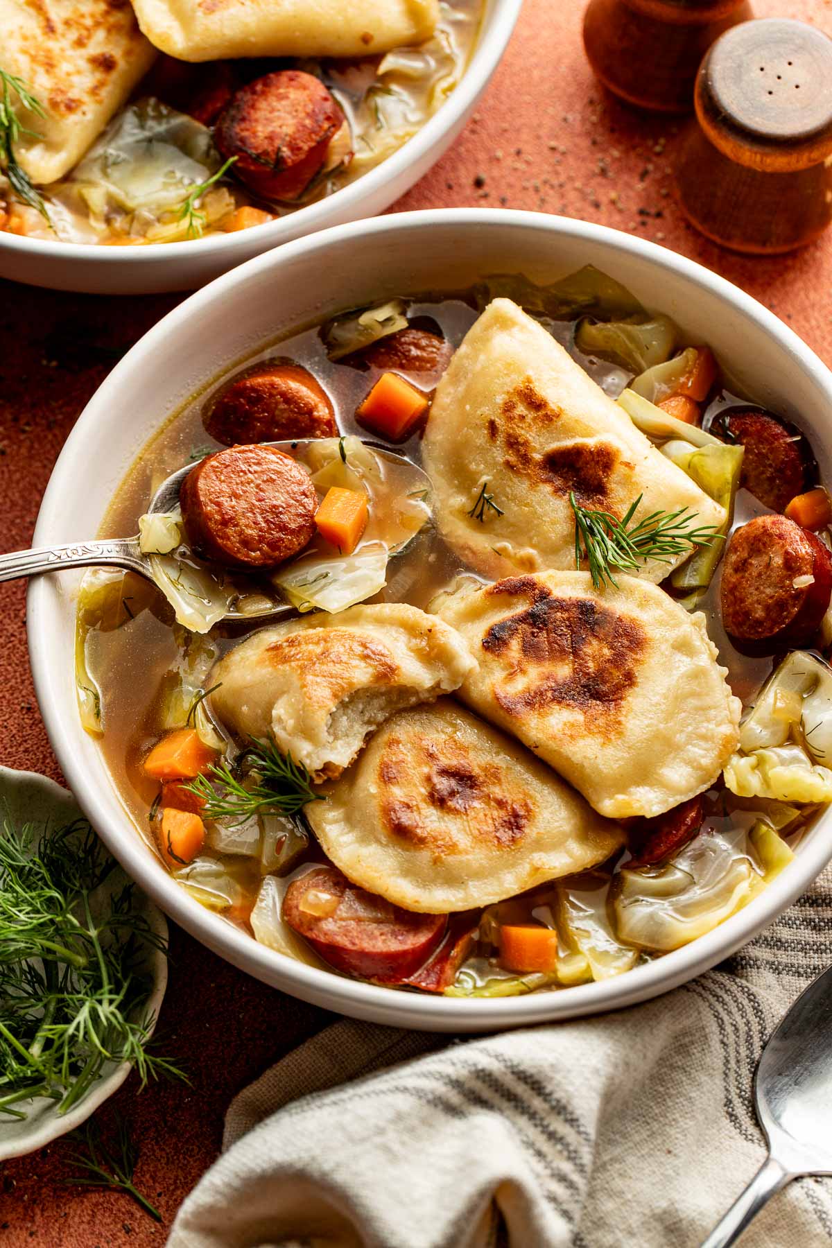 A bowl of pierogi soup featuring pan-fried pierogi, sliced kielbasa, carrots, and cabbage garnished with dill.