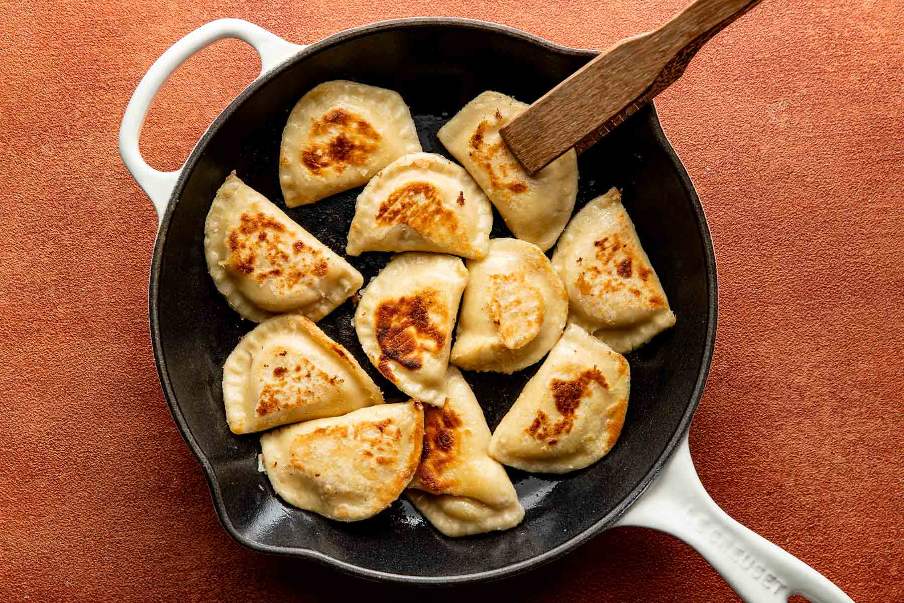 Pan-fried pierogi in a cast iron skillet being turned with a wooden spatula.