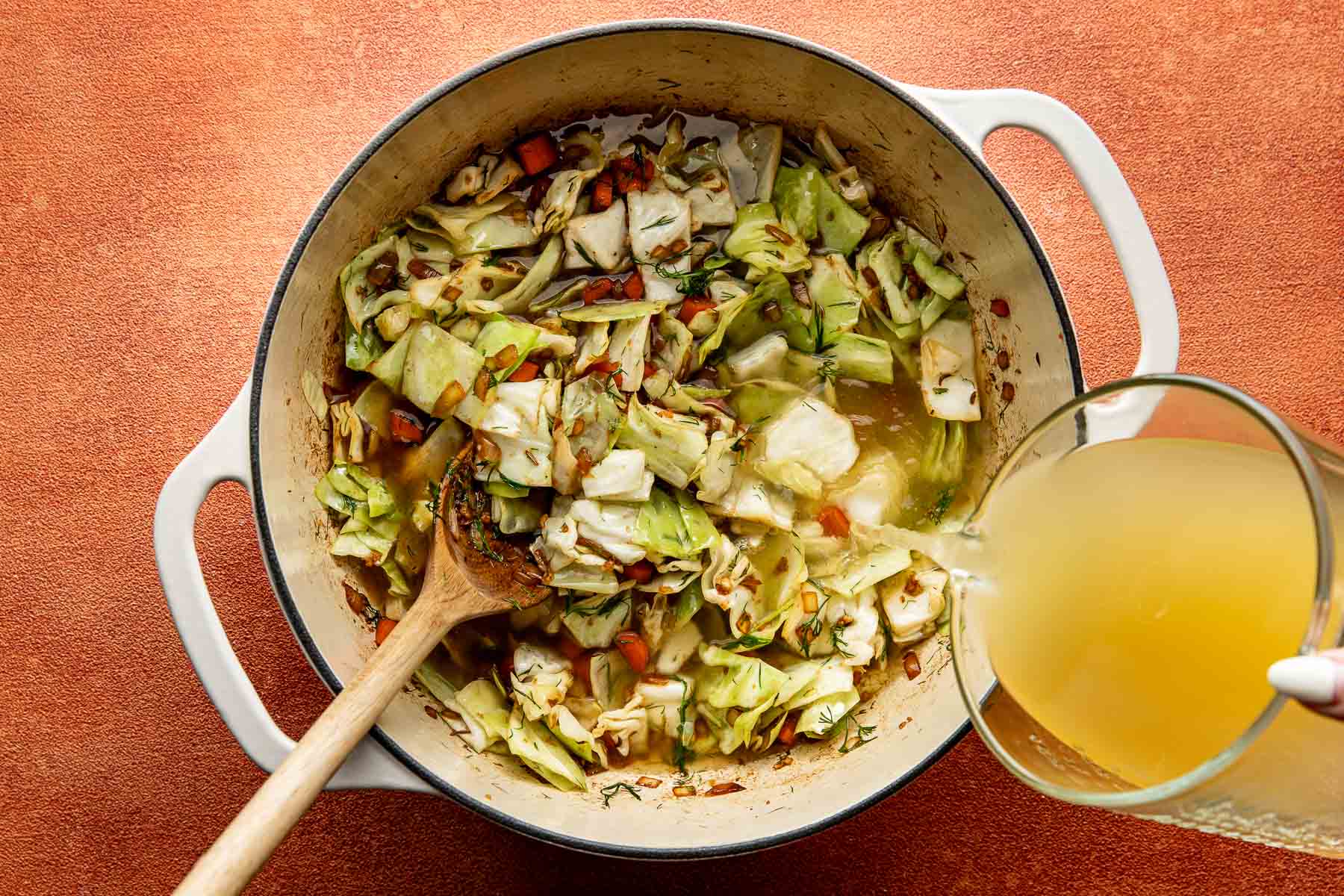Broth being poured into a pot of sautéed cabbage, carrots and onions.