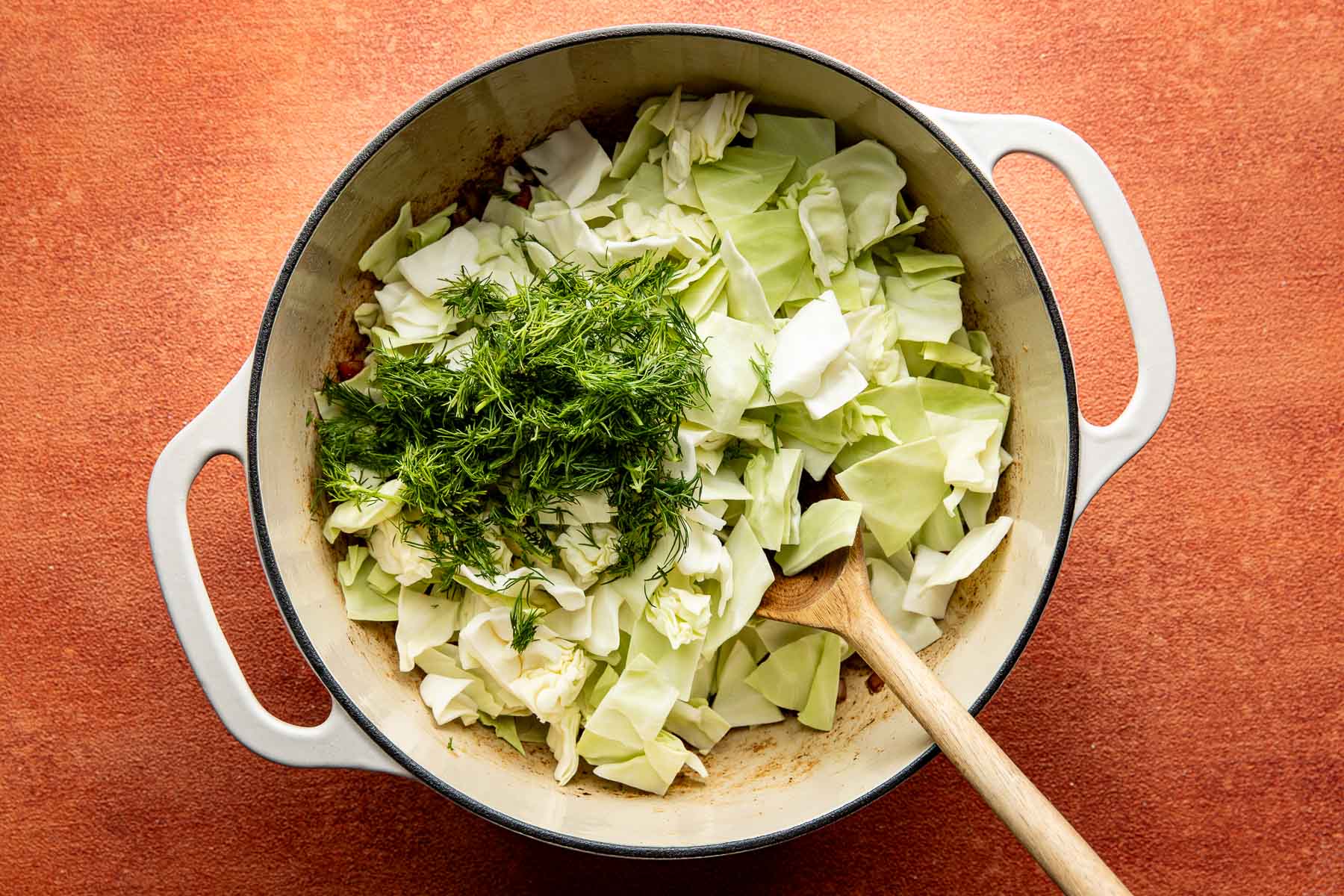 Chopped cabbage and fresh dill in a white pot with a wooden spoon.