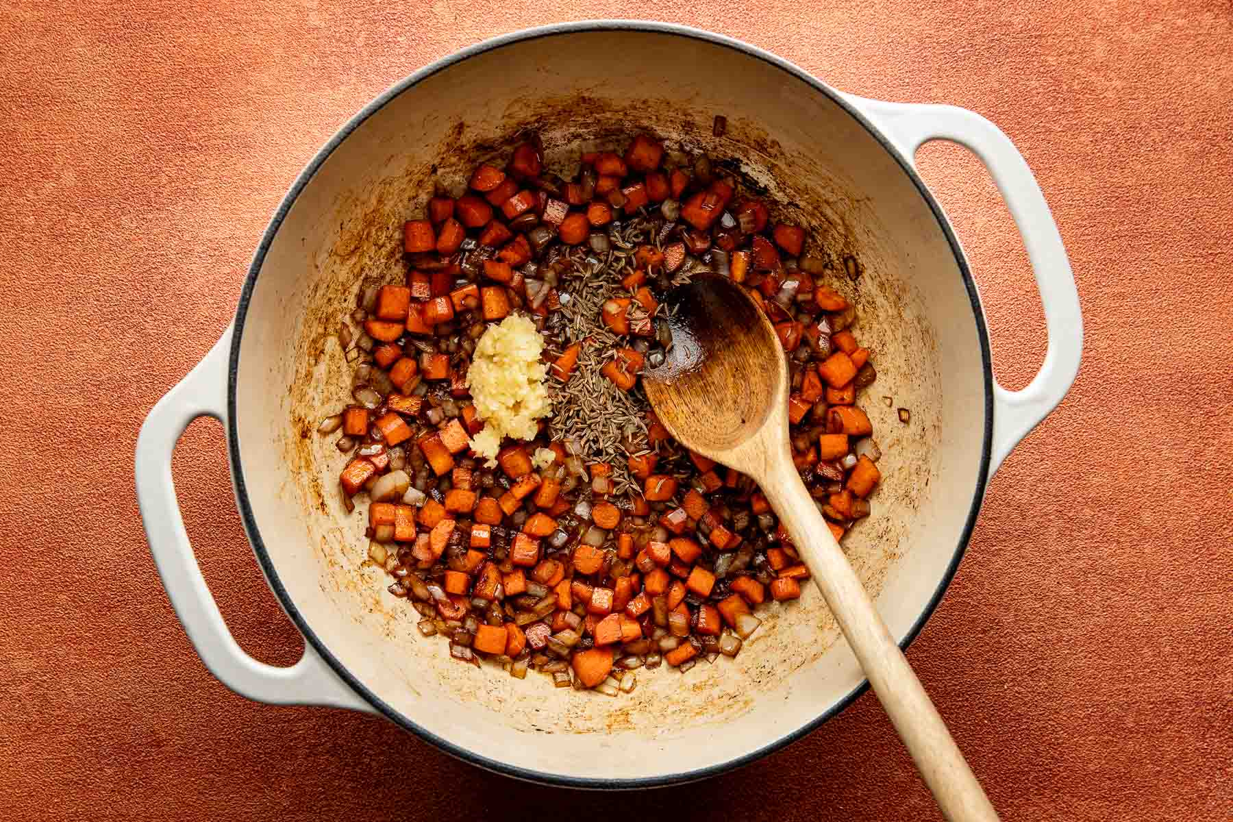 Dutch oven with sautéed carrots and onions, topped with minced garlic and caraway seeds, and a wooden spoon.