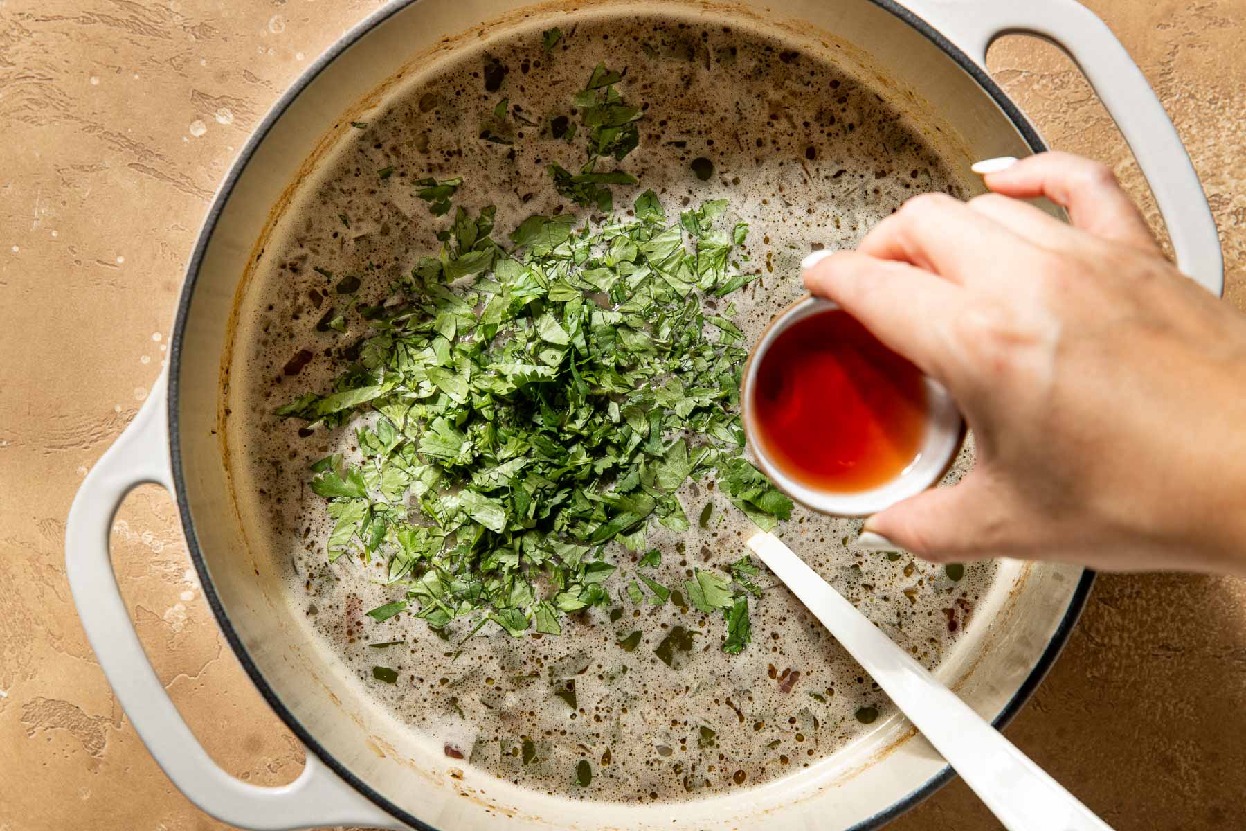 A hand pouring red wine vinegar liquid into a pot of black bean soup with chopped cilantro.