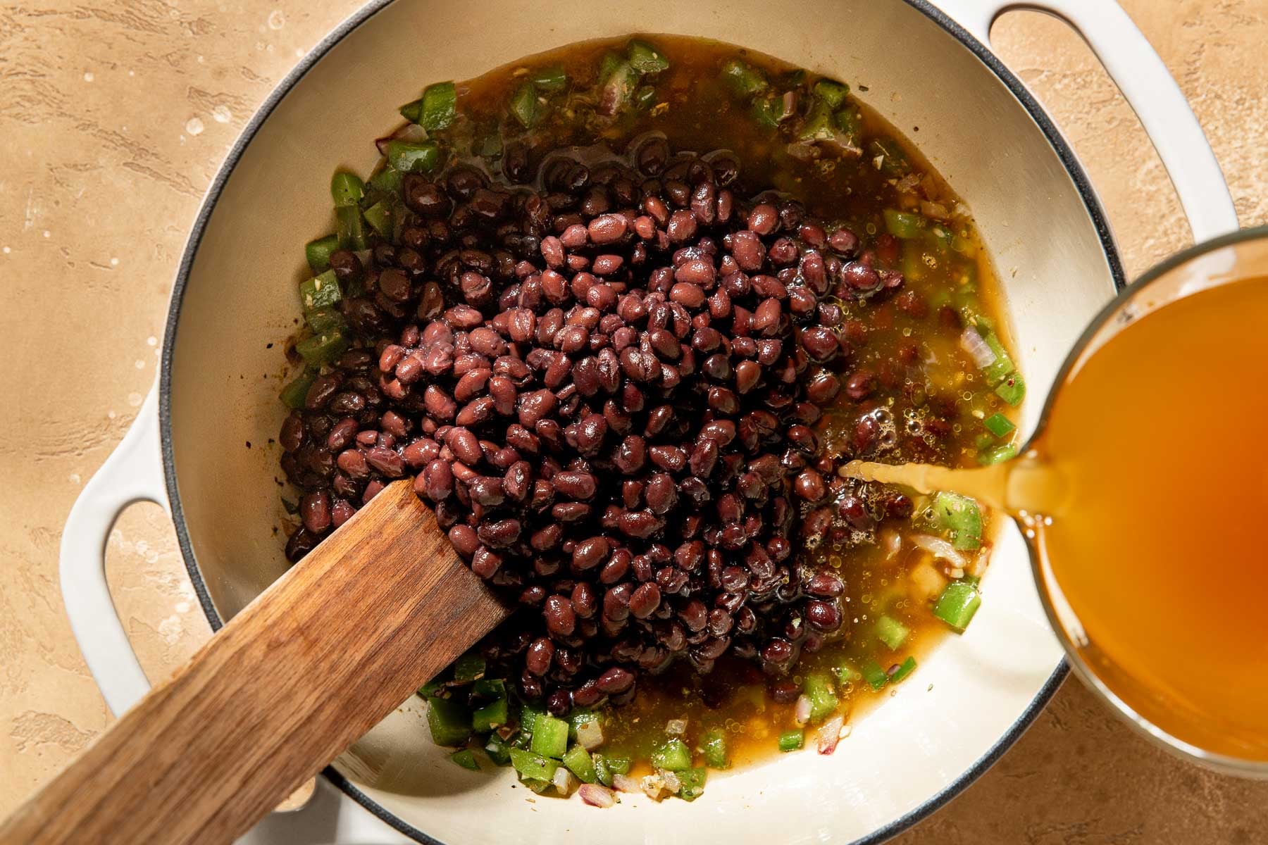 Liquid being poured into a pot containing black beans, chopped peppers, and a wooden spoon.