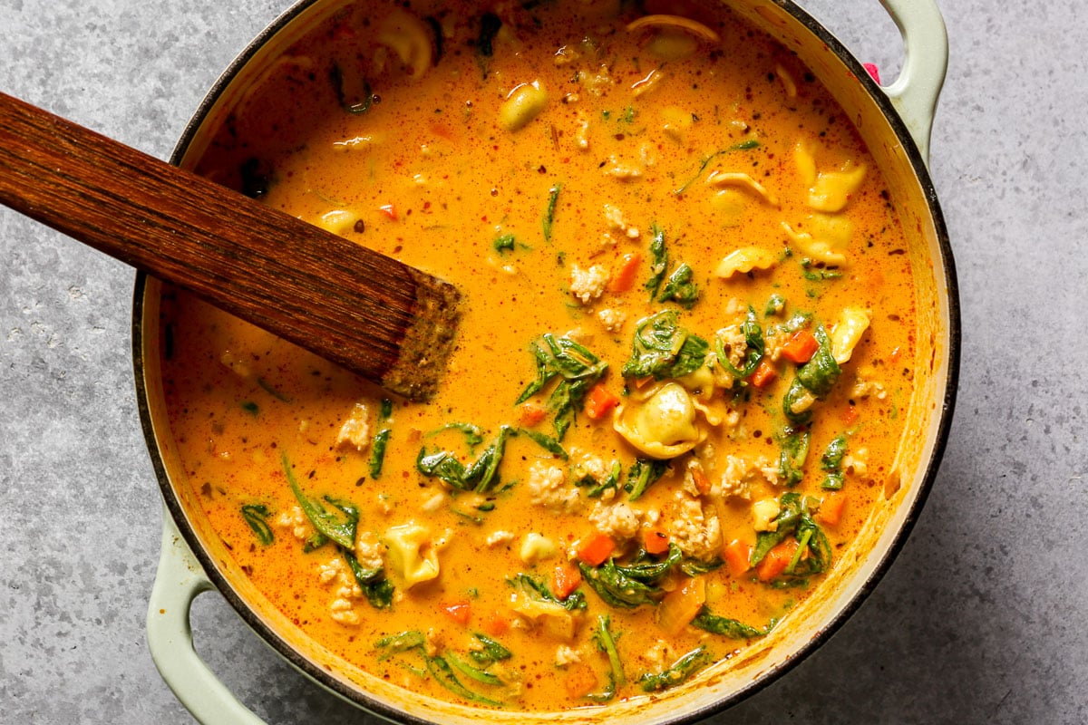 Creamy tomato soup with tortellini, spinach, and meat in a pot with a wooden spoon.