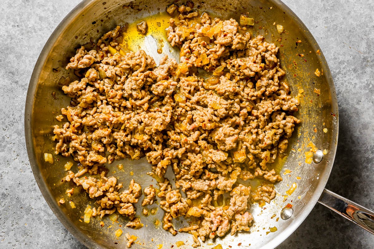 Ground pork browning in a metal skillet with cooked onions.