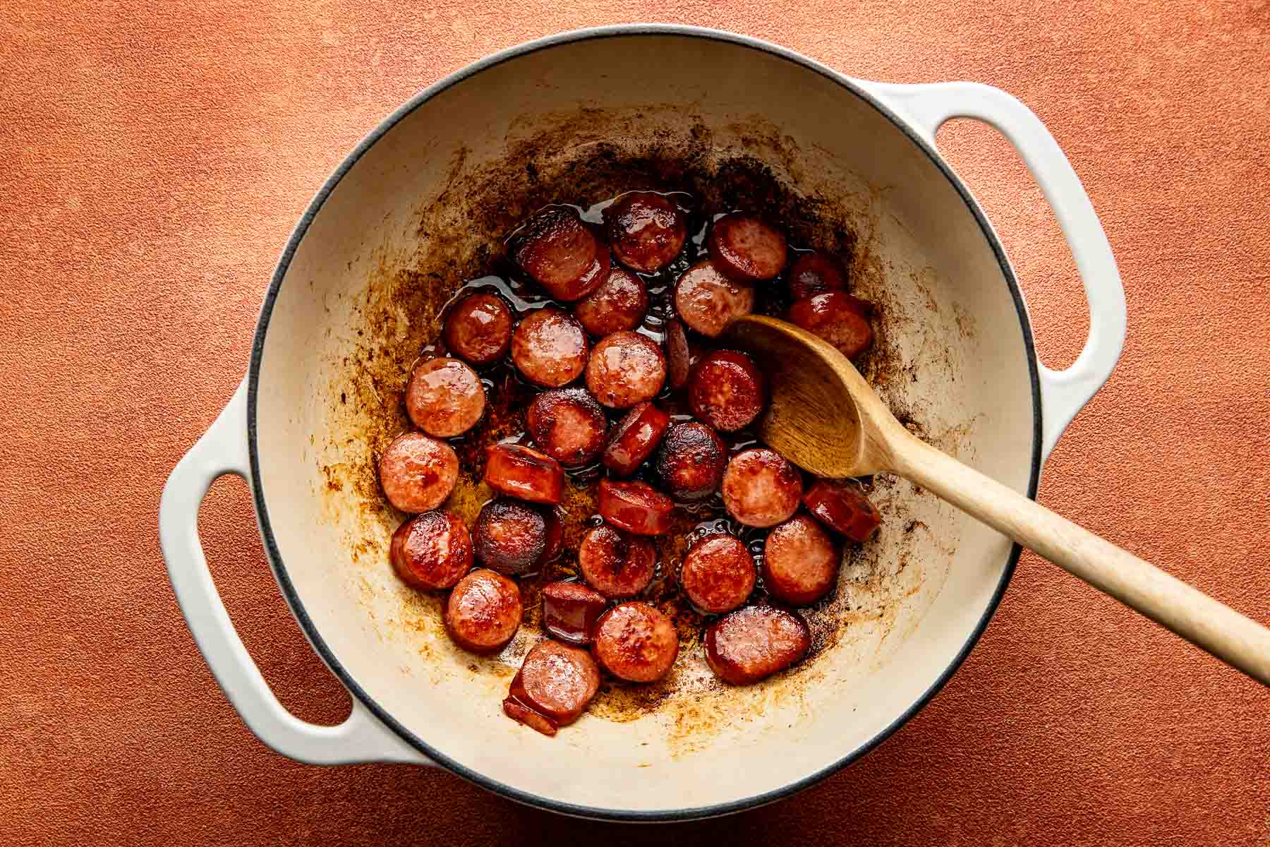 Sliced sausage browning in a white enameled cast iron pot with a wooden spoon.