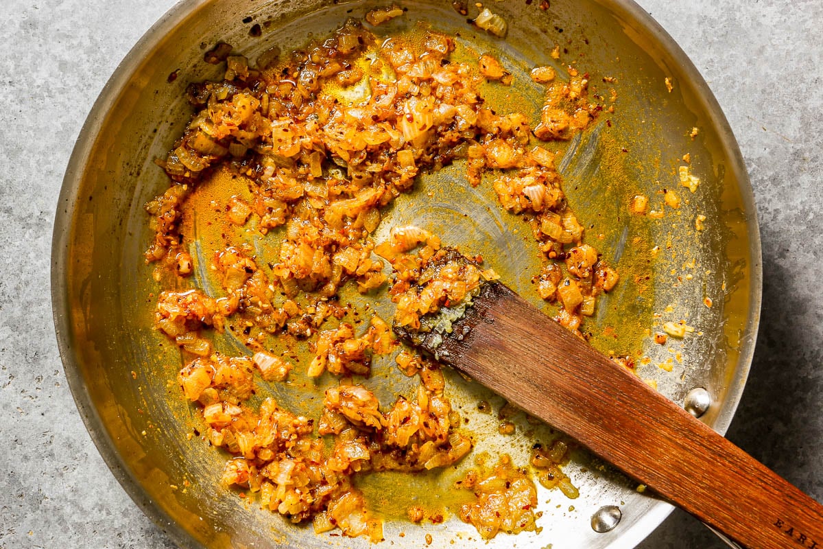 A metal skillet containing sautéed onions and chili crisp being stirred with a wooden spatula.