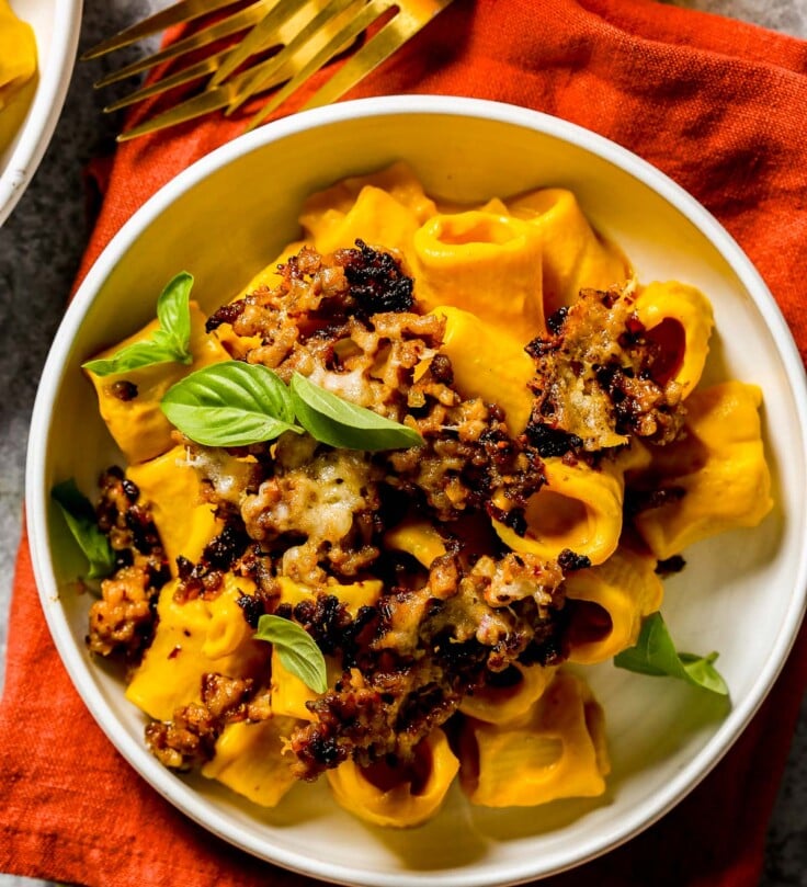 Rigatoni pasta coated in a pumpkin pasta sauce and topping with crispy pork sausage and basil in a white bowl on a red napkin.