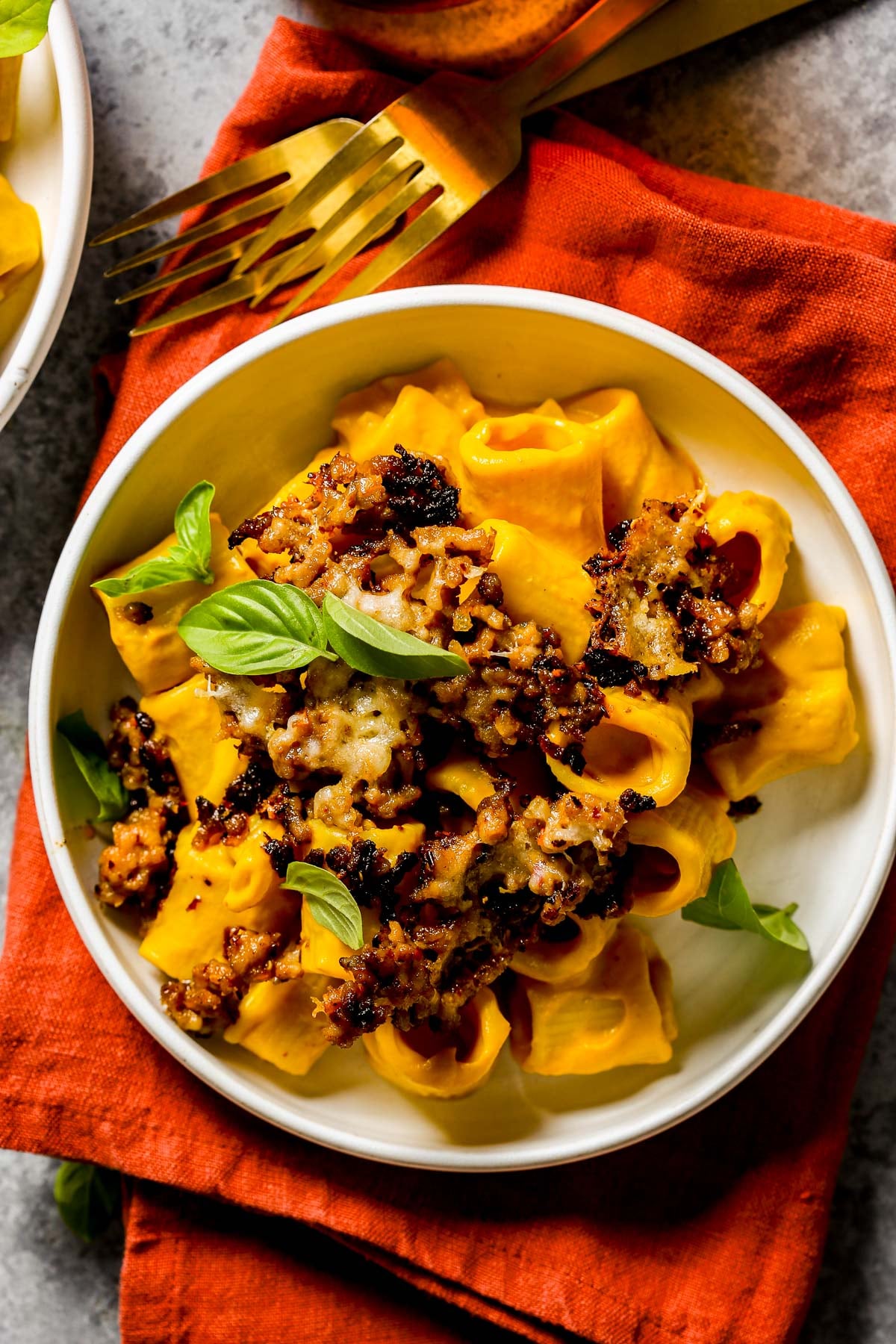Rigatoni pasta coated in a pumpkin pasta sauce and topping with crispy pork sausage and basil in a white bowl on a red napkin.