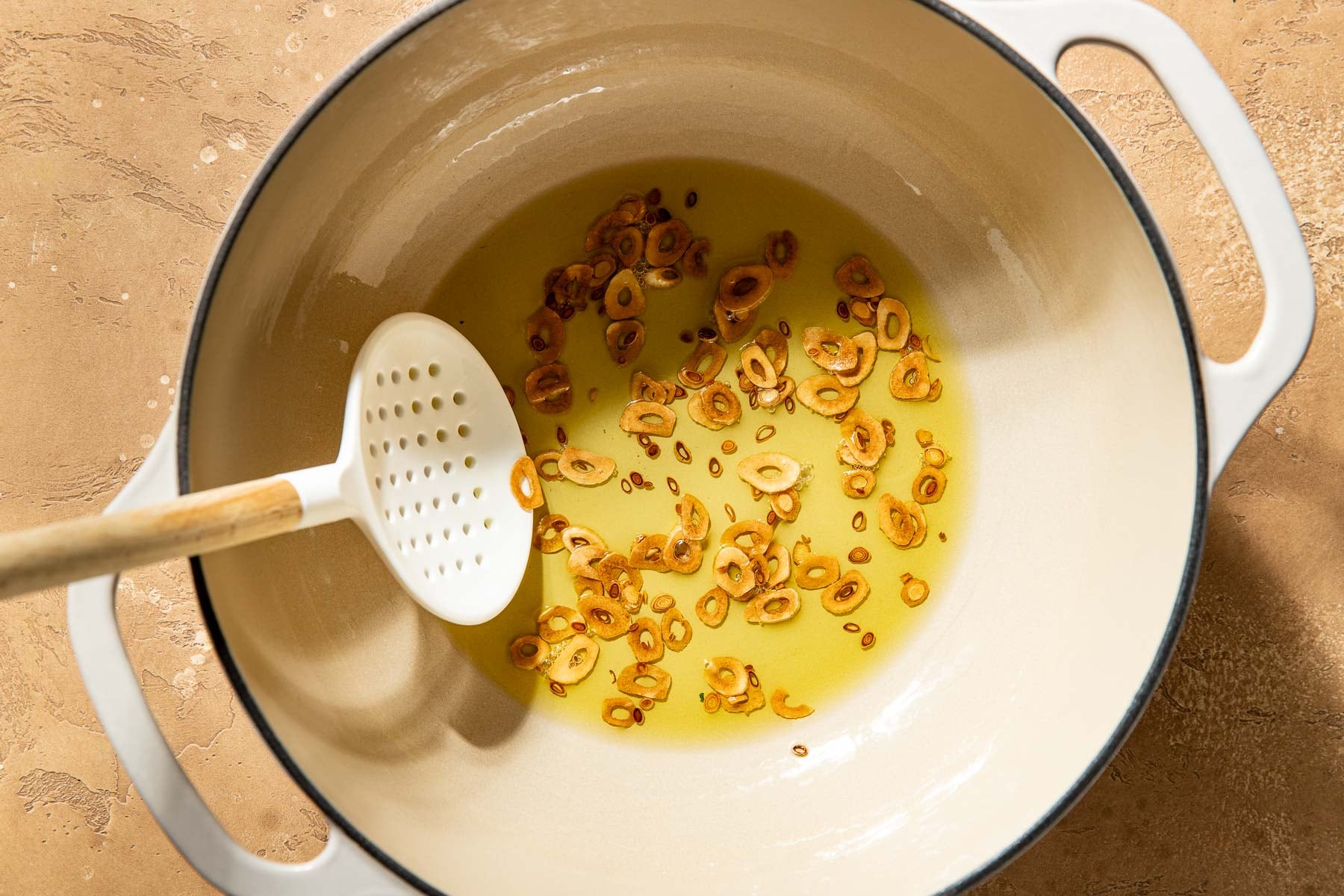 Sliced garlic frying in oil inside a Dutch oven, stirred with a slotted spoon.