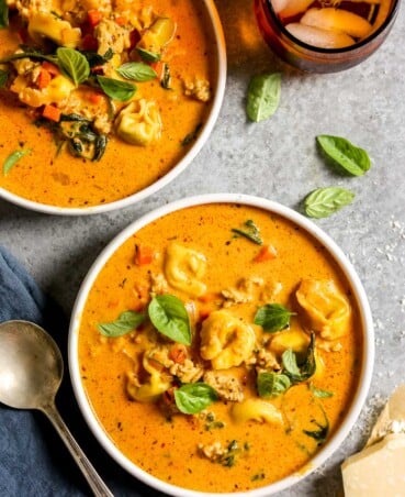 Two bowls of creamy tomato tortellini soup topped with fresh basil, next to a spoon and a glass of iced tea.