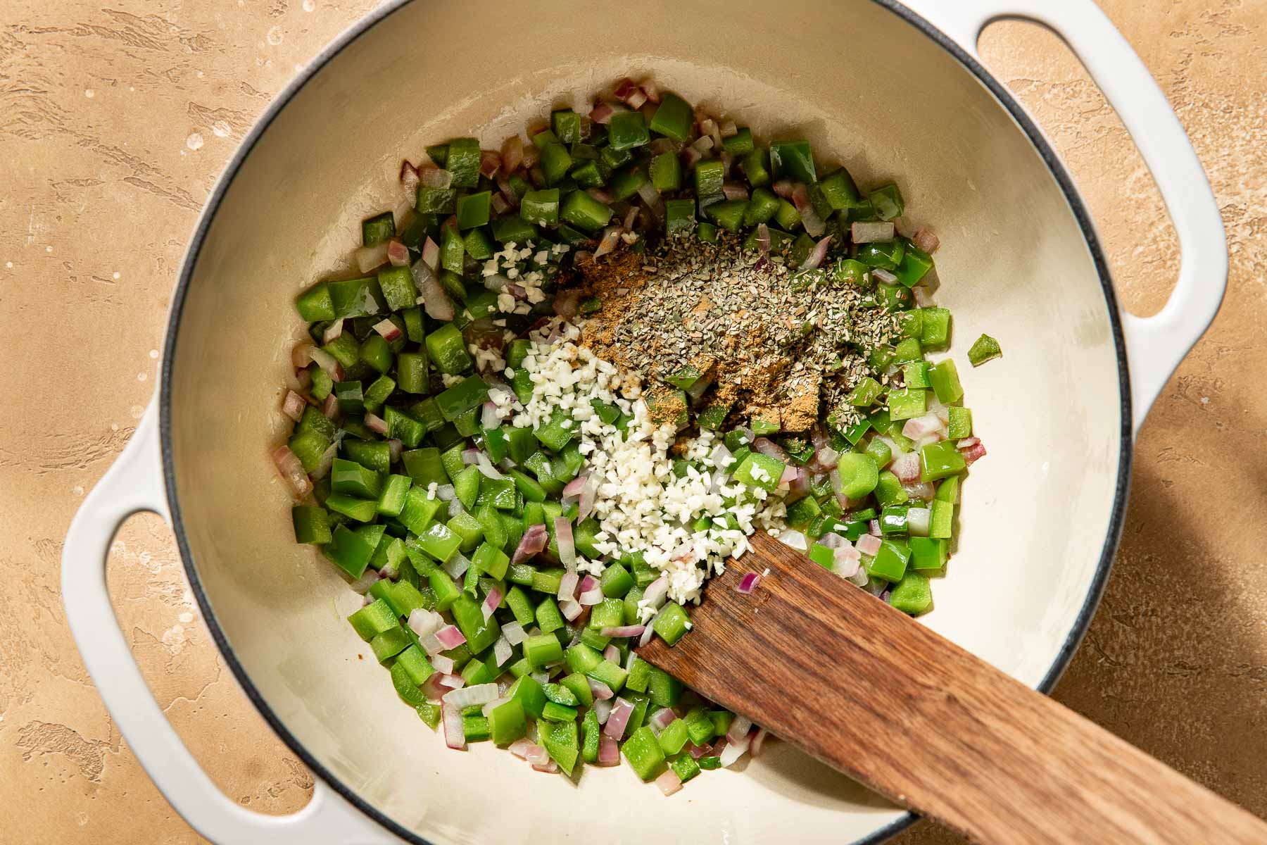 Diced green bell peppers, red onions, garlic, cumin and dried oregano in a white enameled pot with a wooden spatula.
