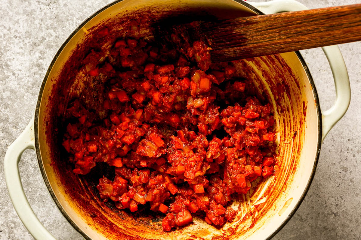A wooden spoon stirs a red, chunky tomato-paste mixture in a large enameled pot.