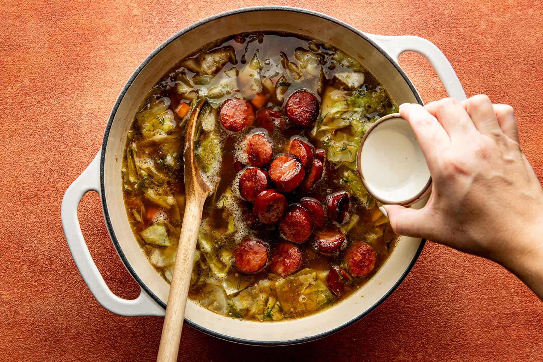 A hand pours vinegar into a pot of cabbage and sliced sausage stew with a wooden spoon.